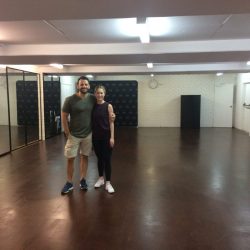 A man and woman stand in an empty dance studio with sprung flooring, ready to begin their practice session.